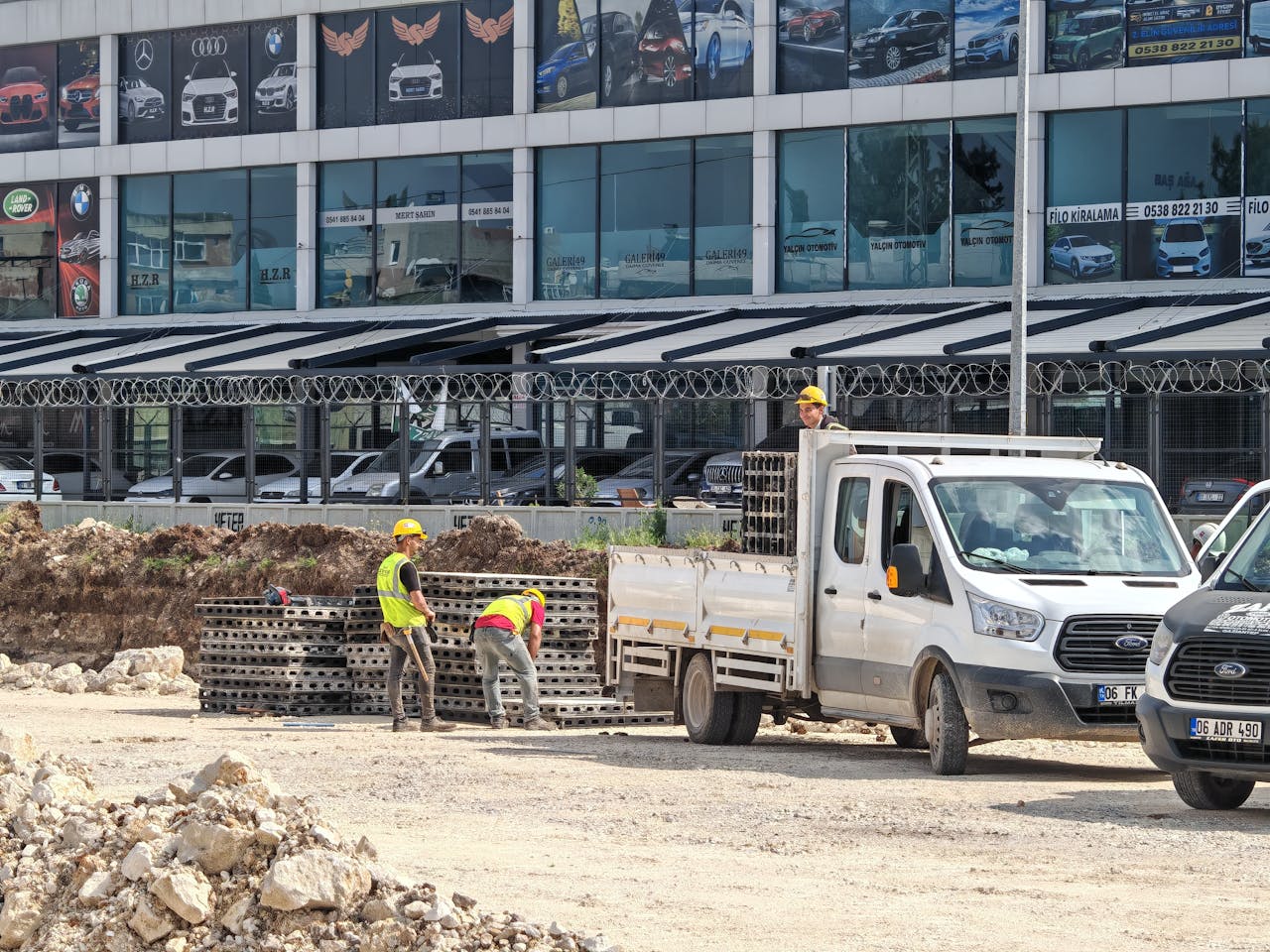 Workers unloading materials from a truck at a construction site in front of a car dealership.